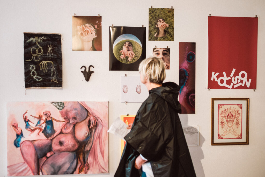 En person med svart jacka och kort blont hår ser på en vägg med olika konstverk. Vi ser henne bakifrån. På väggen hänger en eklektisk blandning av tryck i olika färger. 